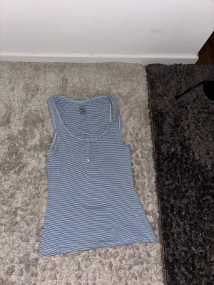 Randig blå linnetopp från Twilfit - Randigt linne med knappar framtill. Lite längre modell och slutar nedanför midjan på ett par lågmidjade byxor.