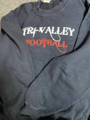 Svart Gildan sweatshirt Tri-Valley - Svart sweatshirt från Gildan med trycket 'Tri-Valley Football' i vitt och rött på bröstet. Tröjan är i modellen Heavy Blend och har rund hals samt långa ärmar. Materialet är en blandning av bomull och polyester, vilket gör den mjuk och skön.