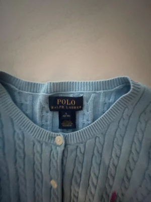 Ljusblå kofta från Polo Ralph Lauren - Snygg ljusblå kofta från Polo Ralph Lauren med kabelstickat mönster och rund hals. Tröjan har långa ärmar, vita knappar framtill och en rosa broderad logga på bröstet. Perfekt för dig som gillar klassisk och stilren design.