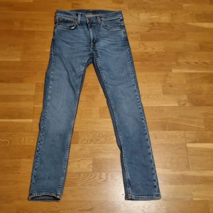 Blå jeans från Nudie Jeans, 29/32 - Säljer ett par klassiska blå jeans från Nudie Jeans i storlek 29/32. Modellen har raka ben, fem fickor och snygga kontrastsömmar. Jeansen är tillverkade i slitstarkt denim och har Nudie Jeans logga på bakfickan. Perfekt passform för dig som gillar en tidlös look.