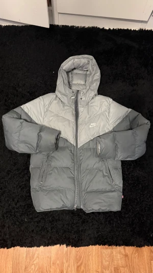 Grå Nike pufferjacka med huva - Säljer en grå pufferjacka från Nike med två nyanser av grått i snyggt blockmönster. Jackan har huva med snörning, dragkedja framtill och två sidofickor med dragkedja. Perfekt för kalla dagar och har en riktigt chill vibe. Helt ny men utan prislapp eller originala förpackningen därför sänktes priset men helt ny kostar 2600