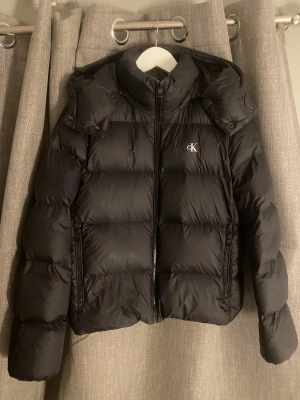 Svart vinterjacka  från Calvin Klein - Svart vinter jacka  från Calvin Klein. Den är använd 2-3 gånger så nästan helt ny. Köpte för 1500kr. Den har två stora fickor framtill och en stor luva bak. Det är storlek M men passar även S. Man fryser inte i den så perfekt nu till vintern 