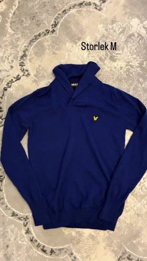 Blå tröja från Lyle & Scott, M - Snygg marinblå tröja från Lyle & Scott i storlek M med gul logga på bröstet. Tröjan har en unik krage och långa ärmar, perfekt för dig som gillar stilrena och klassiska plagg. Materialet känns mjukt och skönt mot huden.