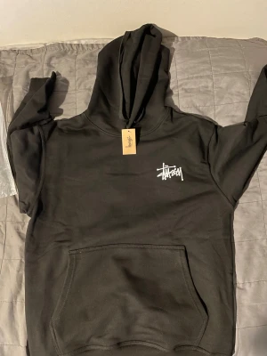 Svart Stüssy hoodie med stor logga bak - Svart hoodie från Stüssy i bomull med stor vit logga på ryggen och mindre logga på bröstet. Klassisk känguruficka framtill och huva med snörning. Perfekt för dig som gillar streetwear och vill ha en clean look.