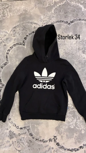 Svart Adidas hoodie med logga - Svart hoodie från Adidas med stor vit logga på bröstet. Tröjan har huva, lång ärm och en klassisk magficka. Perfekt för dig som gillar sportig och avslappnad stil. Materialet är mjukt och skönt, troligen bomull och polyester.