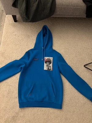 Blå hoodie Supply & Demand NYC i storlek S - Blå hoodie från Supply & Demand med coolt NYC-tryck på bröstet och röd zigzag-söm. Har huva med snörning, känguruficka och lång ärm. Materialet är mjuk bomull och polyester, perfekt för chill dagar. Snygga detaljer och streetstyle-vibe.