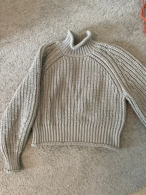 Grå stickad polotröja från H&M - Säljer en chunky grå stickad polotröja från H&M i storlek S. Tröjan har grov ribbstickning, hög polokrage och långa ärmar med breda muddar. Perfekt för dig som gillar en cozy och trendig look under kyliga dagar.