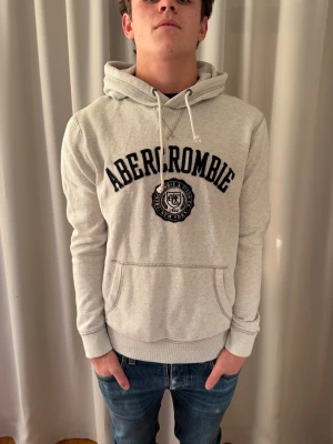 Ljusgrå hoodie Abercrombie & Fitch - Säljer en ljusgrå hoodie från Abercrombie & Fitch med broderad logga och text i mörkblått på bröstet. Tröjan har huva med snörning, känguruficka och ribbade muddar. Perfekt för chill dagar och enkel att matcha med jeans.