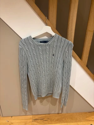 Ljusgrå stickad tröja Polo Ralph Lauren - Snygg ljusblå stickad tröja från Polo Ralph Lauren med klassiskt kabelstickat mönster och rund hals. Tröjan har långärmad passform och en liten broderad logga på bröstet. Perfekt för dig som gillar stilrena och tidlösa plagg.