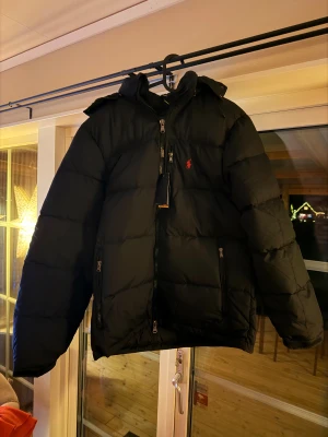 Svart pufferjacka från Polo Ralph Lauren - Svart pufferjacka från Polo Ralph Lauren med röd logga på bröstet. Jackan har huva, flera dragkedjor och quiltad design. Perfekt för kalla dagar och har en modern, loose passform. Materialet är syntet och dun, vilket gör den både varm och lätt. Det är en stor M storlek 