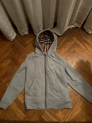 Grå Burberry hoodie med rutig huva - Säljer en grå hoodie från Burberry i storlek M. Tröjan har dragkedja, snörning vid huvan och klassiskt Burberry-rutigt foder i huvan. Broderad Burberry-logga på bröstet och två fickor framtill. Perfekt för dig som gillar stilrena och lyxiga detaljer.