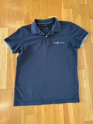 Marinblå pikétröja från Sail Racing - Klassisk marinblå pikétröja från Sail Racing med vit logga på bröstet. Tröjan har krage, knappar framtill och korta ärmar med vita detaljer vid ärmsluten. Perfekt till en avslappnad men stilren look.