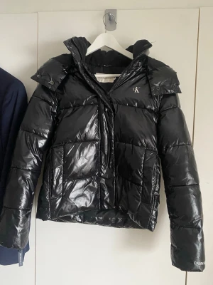 Svart pufferjacka från Calvin Klein - Snygg svart pufferjacka från Calvin Klein med glansig finish och quiltad design. Jackan har huva, dragkedja framtill och CK-logga på bröstet. Perfekt för kalla dagar och ger en cool streetwear-look.