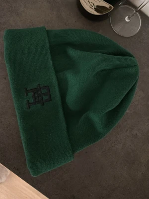 Grön mössa från Tommy Hilfiger - Snygg grön mössa från Tommy Hilfiger med broderad logga framtill. Klassisk modell med uppvikt kant och mjukt, stretchigt material som sitter skönt på huvudet. Perfekt accessoar för att hålla värmen och addera stilpoäng till din outfit💕