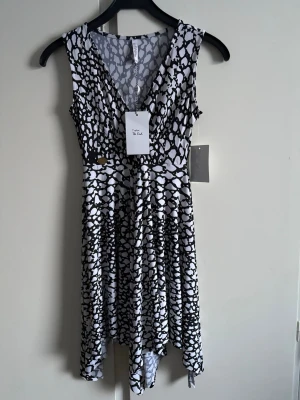 Svartvit mönstrad ärmlös klänning - Snygg ärmlös klänning med svartvitt abstrakt mönster och v-ringning. Klänningen har en asymmetrisk nederkant och är tillverkad i ett mjukt, stretchigt material. Ny med lapp
