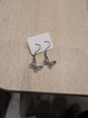 Silvriga fjärilsörhängen i rostfritt stål  - Snygga örhängen i silverfärgad metall med hängande fjärilar. Fjärilarna har en öppen, detaljrik design som ger en lätt och luftig känsla. Perfekta för dig som gillar söta och trendiga accessoarer med naturinspirerat motiv.