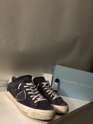 Mörkblå Philippe Model sneakers - Snygga mörkblå sneakers från Philippe Model med vita detaljer och snörning. Skorna har en klassisk låg siluett, vit sula och är tillverkade i mocka med läderdetaljer. Perfekta för dig som gillar stilrena och trendiga sneakers. Skorna i säg är i gott skick men rekommenderar att byta ut orginalsnörenea då dem är extremt slitna som ni ser på bilden.