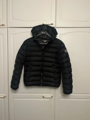 Svart dunjacka från Colmar - Snygg svart dunjacka från Colmar med huva och dragkedja framtill. Jackan har två fickor med dragkedja och en logotyp på ärmen. Perfekt för kalla dagar och enkel att matcha med olika outfits. Jackan är i fint skick. Storlek 14y 166cm passar xs/xxs
