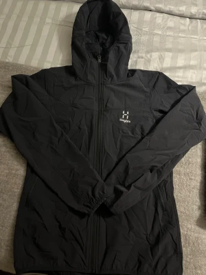 Svart vindjacka från Haglöfs dam - Snygg svart vindjacka från Haglöfs för dam. Jackan har huva, hel dragkedja framtill och två sidofickor med dragkedja. Tillverkad i lätt och slitstarkt material, perfekt för blåsiga dagar. Diskret Haglöfs-logga på bröstet.