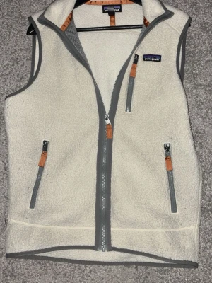 Vit fleeceväst från Patagonia - Mysig vit fleeceväst från Patagonia med grå dragkedjor och detaljer i brunt läder. Västen har tre fickor med dragkedja och hög krage. Perfekt för lager-på-lager och outdoor-vibe.