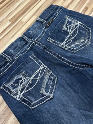 Blå jeans med broderade bakfickor - Snygga blå jeans med unika broderade detaljer och slitningar på bakfickorna. Jeansen har kontrastsömmar och dekorativa nitar som ger en cool look. Passformen är relaxed med raka ben. Säljer då de är för långa för mig. Midjemått: 39cm tvärsöver, innerbenslängden: 83cm. Pris går alltid att diskutera, skriv för fler bilder💝💝