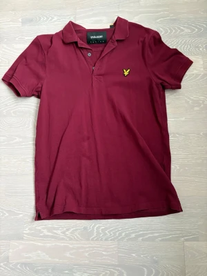 Vinröd pikétröja från Lyle & Scott - Vinröd pikétröja från Lyle & Scott med klassisk krage och knappslå. Tröjan har korta ärmar och den ikoniska gula loggan broderad på bröstet. Perfekt för en stilren och avslappnad look.
