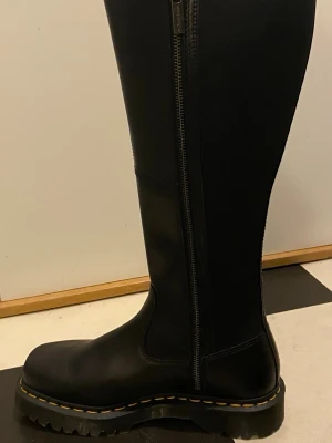 Dr Martens Amaahya Hi Black - Svarta höga stövlar med dragkedja i skinn med grov sula och gul söm längs kanten. Stövlarna har dragkedja på insidan för enkel på- och avtagning. Perfekta för dig som vill ha en cool och robust look. Använda ngr timmar vid ett tillfälle. Nypris 2600
