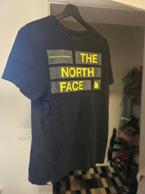 Svart t-shirt från The North Face - Snygg svart t-shirt från The North Face med stort gult tryck på bröstet där det står 'Never Stop Exploring' och loggan. Klassisk passform och rund hals. Perfekt för dig som gillar streetwear och vill ha en enkel men stilren look. 