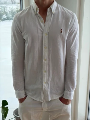 Vit skjorta - Ralph Lauren - Klassisk vit skjorta från Ralph Lauren med button-down krage och broderad logga på bröstet. Skjortan har långa ärmar, normal passform och är tillverkad i bomull. Perfekt för en clean och stilren look.