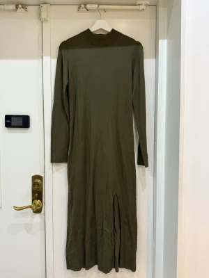 Olivgrön långärmad klänning - En enkel och stilren olivgrön långklänning med lång ärm och hög hals. Klänningen har en rak passform och en slits framtill för extra rörelsefrihet. Perfekt för dig som gillar minimalistisk stil.