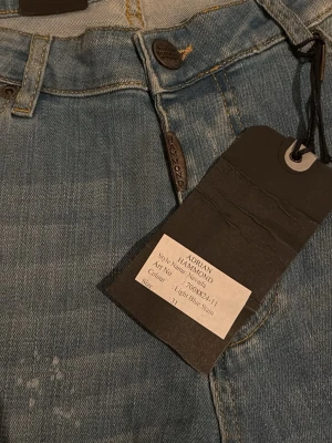 Ljusblå jeans från Adrian Hammond - Snygga ljusblå jeans från Adrian Hammond med slitningar och distressed detaljer. Klassisk femficksmodell med knappgylf och raka ben. Perfekta för en avslappnad och trendig look. Materialet är mjukt och bekvämt jeans i bomull.