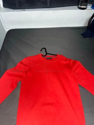 Röd sweatshirt från Sail Racing - Riktigt snygg röd sweatshirt från Sail Racing med broderad logga på bröstet. Tröjan har rund halsringning och ribbade muddar. Perfekt för dig som gillar stilrena och sportiga plagg. Materialet känns mjukt och bekvämt mot huden.