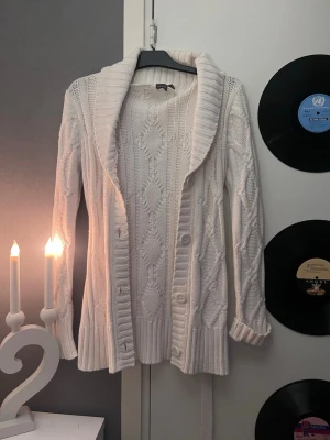 Vit kabelstickad kofta med sjalkrage - Mysig vit kabelstickad kofta med bred sjalkrage och knappar framtill. Koftan har ett bälte i midjan och ribbade muddar vid ärmslut och nederkant. Perfekt för lager på lager och ger en chill men ändå snygg look.