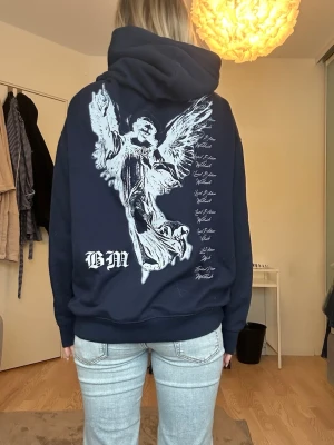 Mörkblå hoodie med ängeltryck - Snygg mörkblå hoodie från BM med stor vit ängel och text på ryggen. Klassisk känguruficka framtill och stor huva. Perfekt för dig som gillar streetwear och vill sticka ut med ett coolt ryggtryck.