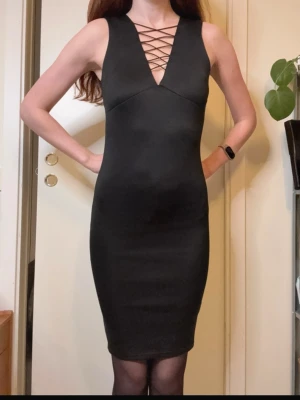 Svart Glansig Tight Figurnära Klänning med Snördetalj - Snygg svart klänning med tight passform och djup v-ringning med snörningsdetalj framtill. Ärmlös modell som slutar ovanför knäna, perfekt för en trendig look. Materialet känns stretchigt och följsamt. Märkt med storlek 8 men skulle säga att det är en XS.
