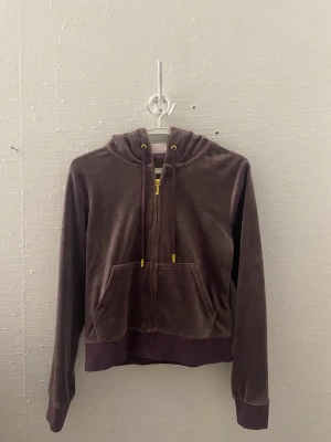 Lila velour hoodie med dragkedja - Mysig lila kofta i mjuk velour från Juicy Couture. Modellen har dragkedja framtill, fickor och huva med snören och guldfärgade detaljer. Ribbstickade muddar vid ärmslut och nederkant. Perfekt för en avslappnad och trendig look.