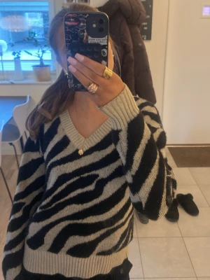 Zebramönstrad stickad tröja. Lappen med storlek och varumärke är bortklippt. - Säljer en stickad tröja med zebramönster i svart och beige. Tröjan har v-ringning, långa ärmar och breda ribbade muddar vid ärmslut och nederkant. Perfekt för dig som vill ha en trendig och mysig look.