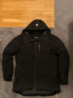 Svart dunjacka Sail Antarctica sail racing  - Svart dunjacka från Sail Antarctica med huva och dragkedja framtill. Jackan har flera fickor med dragkedja och en patch på ärmen. Jackan har används ett bra tag så det finns små defekter  men inga allvarliga, annars är den i fint skick! (Defekter Avdraget från pris) 