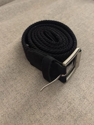 Svart flätat tygbälte med metallspänne - Snyggt svart bälte i flätad textil med ett stilrent silverfärgat metallspänne. Bältet har en flexibel och justerbar design som gör det enkelt att anpassa till olika outfits. Perfekt accessoar för att lyfta din look.