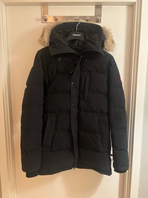 Canada Goose Carson Parka - Svart parkajacka från Canada Goose med pälskantad huva och klassisk logga på ärmen. Jackan är quiltad och har dragkedja samt knappar framtill. Perfekt för kalla dagar med sin varma och rejäla design. Köpt hos Johnells för 10.999kr för några vintrar sedan. Förekommer med både box och papper/kvitto. 