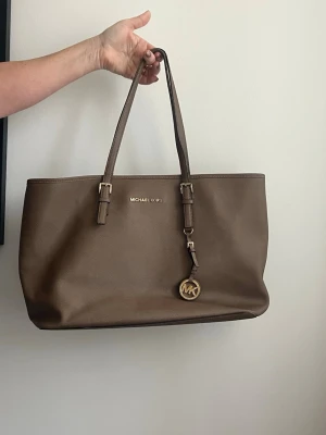 Brun väska från Michael Kors - Brun shoppingväska från Michael Kors i slätt skinn med guldfärgade detaljer och justerbara axelband. Väskan har ett klassiskt MK-hänge och gott om plats för allt du behöver. 