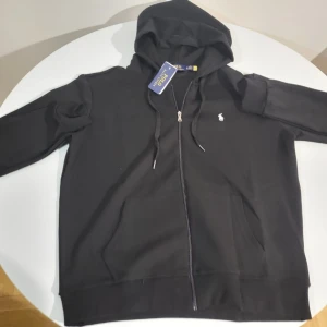 Svart hoodie från Polo Ralph Lauren - Säljer en svart hoodie med dragkedja från Polo Ralph Lauren. Klassisk design med huva, dragsnören och fickor framtill. Liten vit logga broderad på bröstet. Perfekt för en avslappnad och stilren look.