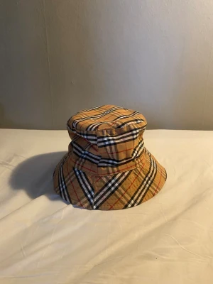 Beige bucket hat med rutigt mönster - Säljer en beige bucket hat från Burberry med det klassiska rutiga mönstret i svart, vitt och rött. Hatten har mjuk form och bred skärm, perfekt för en trendig streetwear-look. Ikonisk accessoar som lyfter vilken outfit som helst.