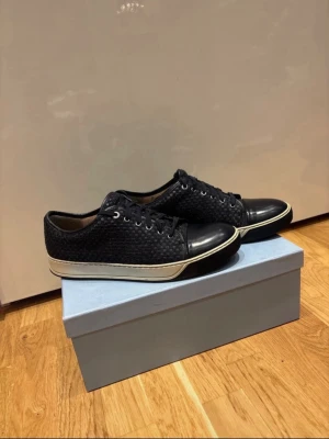 Svarta sneakers från Lanvin - Snygga svarta sneakers från Lanvin med flätad textil på sidorna och tå i skinn. Klassisk låg modell med snörning och vit sula. Perfekta för dig som vill ha en stilren och exklusiv look. Skriv vid frågor och pris kan diskuteras.