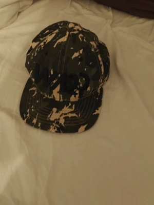 Keps med camomönster och svart text - Säljer en keps från Name It med coolt camouflagemönster i grönt, beige och svart. Framtill står det 'Wapp' i svart text. Kepsen har böjd skärm och justerbar passform. Perfekt för dig som vill sticka ut med en streetstyle-look.