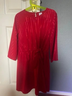 Röd glittrig klänning från Lindex - Röd kortklänning från Lindex med glittriga ränder och trekvartsärm. Klänningen har ett knyt i midjan och rund halsringning.