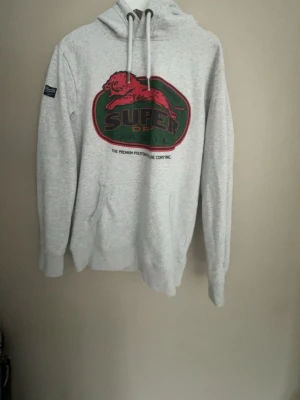 Grå Superdry hoodie med tryck - Snygg ljusgrå hoodie från Superdry med stort rött och grönt tryck på bröstet och en huva med vita dragsnören. Klassisk känguruficka framtill och ribbade muddar. Perfekt för dig som gillar streetwear och vill ha en bekväm och stilren look.