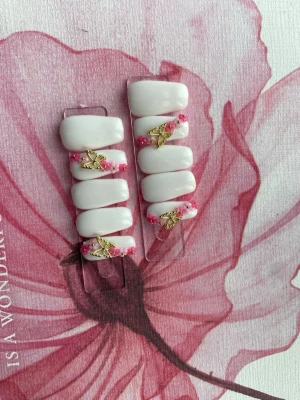 Vita press-on naglar med rosa dekoration - Snygga press-on naglar i vit färg med rosa pärlor och guldfärgade fjärilsdetaljer. Naglarna har en mjukt rundad form och ger en elegant look. Perfekta för dig som vill ha något extra till naglarna. Materialet är plasto gele.