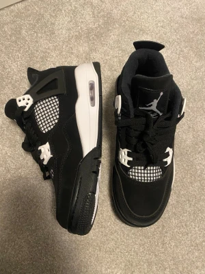 Svarta Nike Jordan 4 sneakers - Snygga svarta Nike Jordan 4 sneakers med vita detaljer och klassisk Jumpman-logga på plösen och hälen. Skorna har snörning och en vit sula med synlig Air-enhet. Perfekta för dig som gillar streetwear och basketskor.