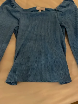Blå långärmad topp från H&M - Säljer en blå långärmad topp från H&M i storlek XS. Toppen har en ribbad struktur och en rak, något croppad passform. Materialet är mjukt och stretchigt, perfekt för att styla med jeans eller kjol. Enkel men snygg design som passar till många looks.